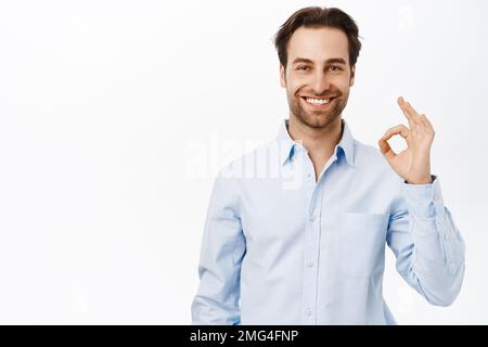 Selbstbewusst lächelnder Geschäftsmann, Verkäufer zeigt okay, OK Zeichen in Genehmigung, wie smth, empfiehlt Unternehmen, steht über weißem Hintergrund Stockfoto
