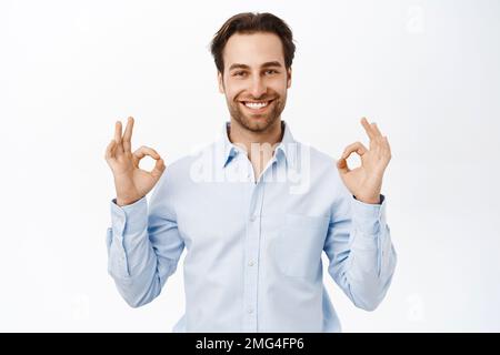 Selbstbewusst lächelnder Geschäftsmann, Verkäufer zeigt okay, OK Zeichen in Genehmigung, wie smth, empfiehlt Unternehmen, steht über weißem Hintergrund Stockfoto