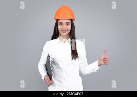 Geschäftsbauerin mit Klemmbrett, Porträt auf Grau. Porträt einer Arbeiterin mit Daumen nach oben im Helm. Stockfoto