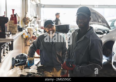 Mittlere Aufnahme. Zwei männliche Mechaniker stehen neben einer Werkbank in einer Werkstatt, lächeln und schauen in die Kamera. Hochwertiges Foto Stockfoto