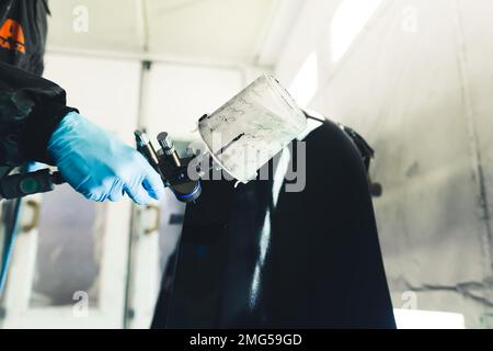 Person trägt Schutzkleidung und Handschuhe, die Airbrush zum Lackieren von schwarzem Auto hält. Autolack. Horizontaler Innenschuss. Hochwertiges Foto Stockfoto