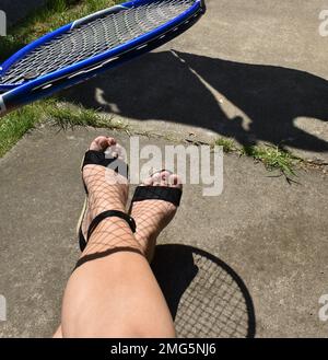 Sommer- und Frauenfüße in schwarzen Sandalen und Schatten des Tennisschlägers als Netzsocken. Stockfoto
