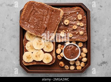 Holzbrett mit leckeren Toasts mit Haselnussbutter, Banane und Nüssen auf hellem Hintergrund Stockfoto