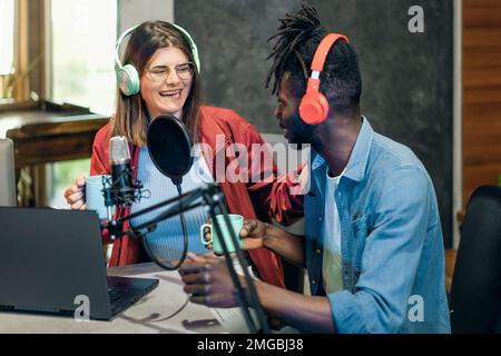 Zwei Content-Ersteller sitzen an einem Tisch und nehmen einen Podcast auf. Der afroamerikanische Mann und die weiße Frau tragen beide Kopfhörer und konzentrieren sich auf ein Tablet Stockfoto