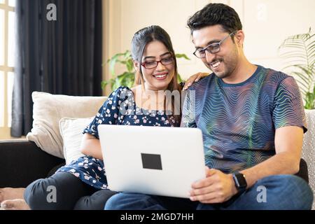Glückliches, lächelndes indisches Paar mit Brille, das mit dem Laptop beschäftigt ist, während es zu Hause auf dem Sofa sitzt - Konzept, die Inneneinrichtung zu überprüfen, online Stockfoto