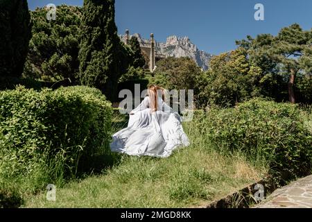 Eine schöne Frau mit langen braunen Haaren und langen weißen Kleid bleiben entlang eines Pfades entlang schönen Büschen in der Park Rückansicht Stockfoto