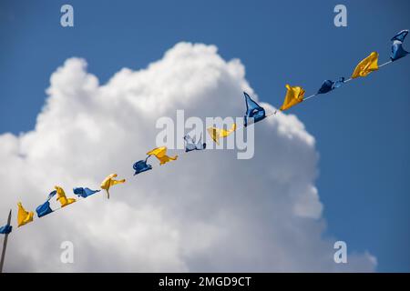 Gelbe und blaue dreieckige Festivalflaggen am Himmel mit weißen Wolken. Feier Im Freien. Festliche Stimmung Stockfoto