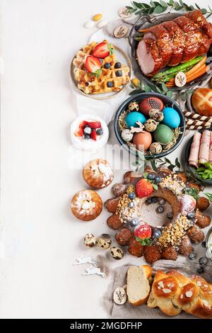 Traditionelles Osterabendessen oder Brunch mit Schinken, bunten Eiern, warmen Brötchen, Kuchen und Gemüse. Ostergerichte mit holtagstauglichen Dekorationen. Draufsicht Stockfoto