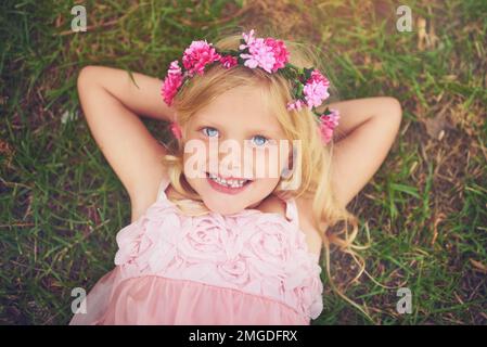 Entspannt in der Sonne. Ein glückliches kleines Mädchen, das in die Kamera schaut, während es auf dem Gras liegt und sich draußen in der Natur entspannt. Stockfoto