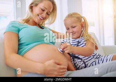 Hören wir, was das Baby sagt... ein kleines Mädchen, das ein Stethoskop benutzt, um zu Hause den schwangeren Bauch seiner Mutter zu hören. Stockfoto