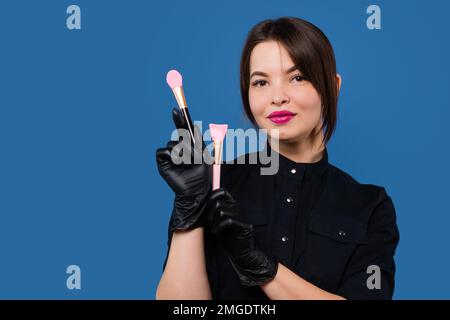 Kosmetiker hält Set Spatel zum Maskieren und Abziehen. Kosmetikerin in schwarzer Uniform und Handschuhen auf blauem Studiohintergrund Stockfoto