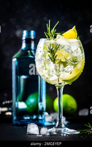 Gin Tonic Cocktail mit trockenem Gin, Rosmarin, Tonic, Limette und Eiswürfeln in Weinglas. Schwarzer Balkenhintergrund, Balkenwerkzeuge Stockfoto