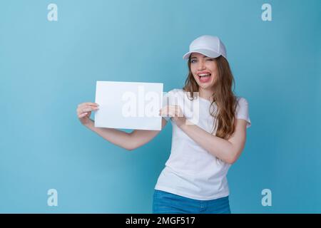 Glückliche Frau, die eine Broschüre mit leerem Einband auf blauem Hintergrund hält und weißes T-Shirt und Kappe für Modell trägt, Draufsicht. DIP für Design Stockfoto