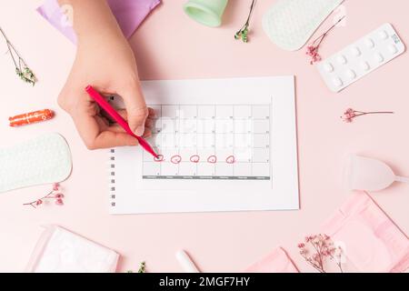 Frauenspuren mit rotem Filzstift im Kalender der Menstruationszyklus auf pinkem Hintergrund mit Pillen und Sanitärserviette Stockfoto