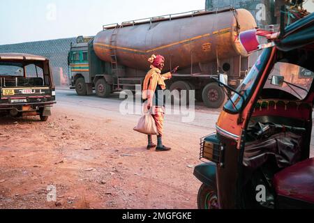 Ein alter Indianer trägt einen roten Turban und orangefarbene Dhoti Kurta mit einer Jacke, die auf der Hauptstraße auf ein Transportfahrzeug wartet, ein alter Mann steht auf der Hauptstraße Stockfoto