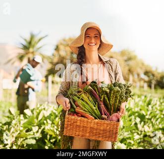 Selbstgebautes Glück. Porträt eines glücklichen jungen Landwirts, der Kräuter und Gemüse in einem Korb auf seinem Hof erntet. Stockfoto