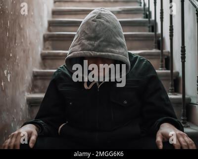 Ein angespannter Mann in der Kapuze, der auf der Treppe sitzt. Drogensucht oder Depressivität. Stockfoto