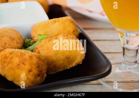 Kroketten oder traditionelle hausgemachte spanische Kroketten auf einem schwarzen Teller mit einem Bier auf der Seite. Konzept für Tapas-Speisen Stockfoto