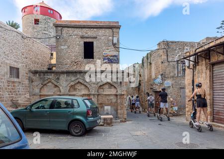 Rhodos, Griechenland - 24. August 2022: Blick auf die Altstadt von Rhodos, Griechenland. Gepflasterte Straßen und Gehwege mit Steinhäusern. Eine Gruppe von Personen macht eine Stadtbesichtigung auf einem dreirädrigen Motorroller. Stockfoto