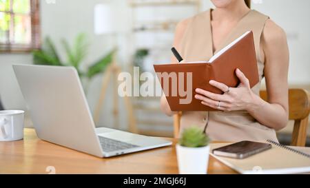 Zugeschnittenes Bild, wunderschöne asiatische Geschäftsfrau der Millennials oder Chefin, die an ihrem Schreibtisch arbeitet, sich Notizen macht oder Listen auf ihrem Notizbuch macht. Stockfoto