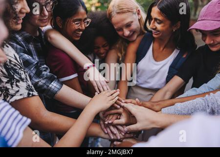 Verschiedene Teenager lächeln fröhlich, während sie ihre Hände in einer Besprechung zusammenlegen. Gruppe von Jugendlichen der Generation z, die Teamgeist und Toga symbolisieren Stockfoto