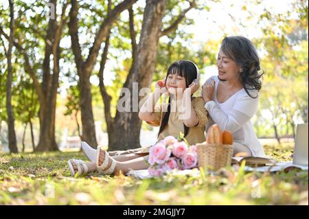 Fröhliche und freundliche asiatische Großmutter, die mit ihrer lieblichen Enkelin im Park picknickt und am Wochenende eine glückliche Familienzeit zusammen hat. Freizeit und f Stockfoto