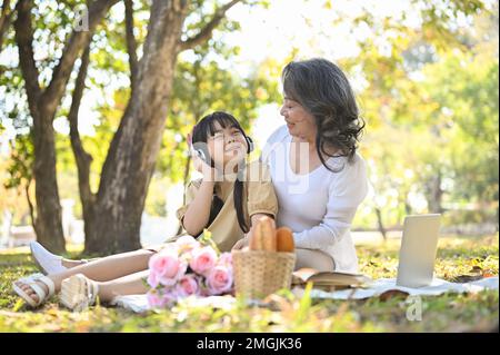 Fröhliche asiatische Großmutter, die mit ihrer lieblichen Enkelin im wunderschönen Park picknickt und am Wochenende eine glückliche Familienzeit hat. Freizeit und Stockfoto