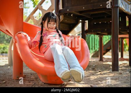 Ein süßes und fröhliches asiatisches Mädchen spielt auf dem Spielplatz, gleitet auf der Rutsche, hat Spaß im Park, genießt Outdoor-Aktivitäten. Stockfoto