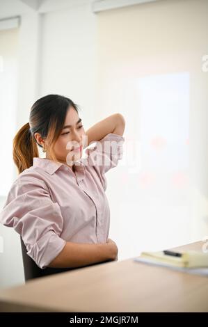 Porträt, müde asiatische Geschäftsfrau oder Büroangestellte, die unter Rückenschmerzen oder Nackenschmerzen leidet und ihren Nacken massiert, während sie an ihr sitzt Stockfoto