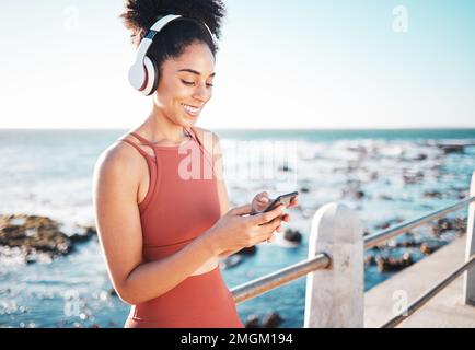 Schwarze Frau am Strand, Kopfhörer und Telefon mit Fitness, Läufer hört Musik für sportliche Motivation. Viel Spaß beim Online-Streaming mit Podcast oder Radio Stockfoto