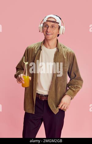 Junger, positiver Mann in stilvollen Kleidern, der Musik über Kopfhörer hört, Orangensaft trinkt, vor pinkfarbenen Studiohintergründen posiert. Emotionsbegriff Stockfoto