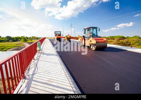 Blick auf mehrere Dampfwalzen, die frischen Asphalt abflachen und eine Schicht heißen Asphalts auf vorbereitetem Boden an der im Bau befindlichen Brücke verteilen. Stockfoto