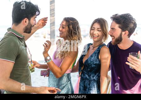 Vier kaukasische Freunde genießen einen Sommertag am Strand und tanzen im Schatten eines chiosco. Die Gruppe lächelt alle, wenn sie loslassen und ein gutes Lachen haben Stockfoto
