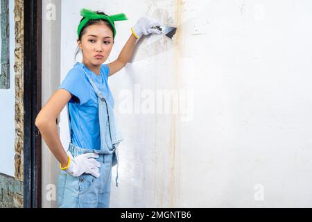 Asiatische junge Hausfrau im Handschuh mit einem Farbschaber an der Wand Stockfoto