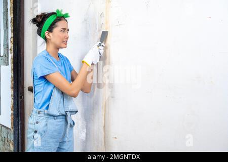 Asiatische junge Hausfrau im Handschuh mit einem Farbschaber an der Wand Stockfoto