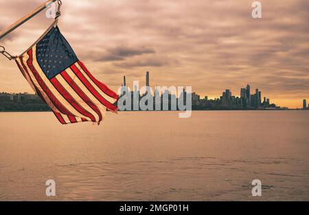 amerikanische Flagge und malerischer Blick auf die Skyline von Manhattan, New York bei Sonnenuntergang vom Hudson River in Edgewater, New Jersey Stockfoto
