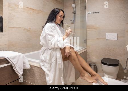 Eine wunderschöne Brünette genießt einen Abend allein im Bad mit einem Glas Prosecco, sitzt auf der Badewanne und ist nur in einem Bademantel gekleidet. Stockfoto