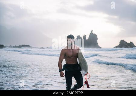Surfer mit Surfbrettspaziergängen am Strand, attraktiver Kerl schaut sich um Stockfoto