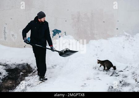 Schneeräumung vor dem Haus. Die Katze sieht zu, wie der Mann den Schnee reinigt. Hochwertiges Foto Stockfoto