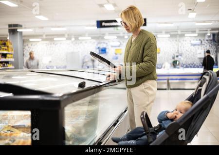 Casualy kleidete Mutter, die Tiefkühlprodukte in der Supermarkt-Abteilung auswählte, zusammen mit ihrem kleinen Jungen im Kinderwagen Stockfoto