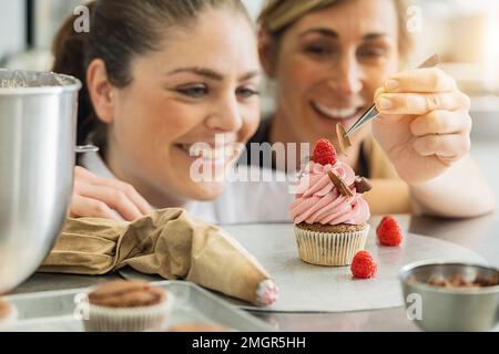 Ein weibliches Team von Konditoren drapierte Schokoladenstücke mit einer Pinzette auf einem Muffin in einer Bäckerei Stockfoto