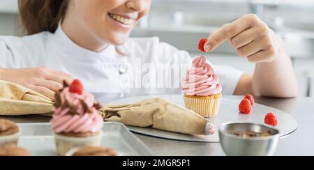 Frauen in der Konditorei, die Muffins machen und eine Himbeere drauf legen Stockfoto