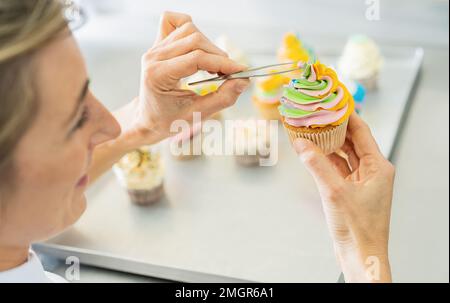 Die Frauen in der Bäckerei, als der Konditor Süßigkeiten mit Perlenverzierung auf einen bunten Muffin mit Pinzette drapierte Stockfoto