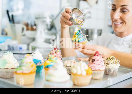 Frauen in der Bäckerei, während der Konditor die Mischung zerbröselt und von Hand auf einen bunten Muffin verzichtet Stockfoto