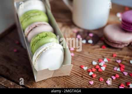 Makronen in einer Schachtel, eine Tasse Kaffee, ein Milchkännchen vor dem Hintergrund kleiner Herzen auf einem hölzernen Hintergrund, Draufsicht Stockfoto