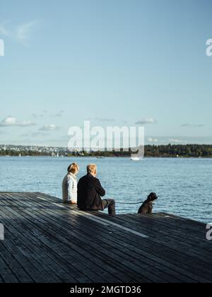 Ein Seniorenpaar mit ihrem Hund, der auf dem hölzernen Dock über dem See unter blauem Himmel sitzt Stockfoto