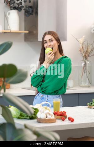 Eine attraktive Brünette hält einen grünen Apfel, während sie am Küchentisch steht. Stockfoto