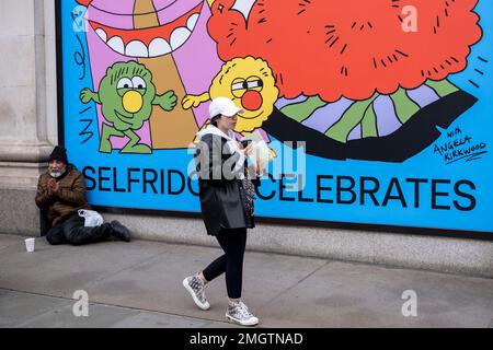 Einkäufer und Besucher passieren eine Schaufensterausstellung mit Illustrationen von Angela Kirkwood Selfridges vor dem berühmten Kaufhaus, wo ein Mann am 9. Januar 2023 in der Oxford Street in London, Großbritannien, um Geld bettelt. Die Oxford Street ist ein bedeutendes Einkaufszentrum im West End der Hauptstadt und mit rund einer halben Million Besuchern täglich die geschäftigste Einkaufsstraße Europas mit rund 300 Geschäften, von denen die meisten Mode- und Bekleidungsgeschäfte sind. Stockfoto