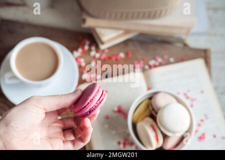 Eine Hand hält eine lila Makrone über einer Tasse Kaffee und einem Buch. stimmungsvolles Foto Stockfoto