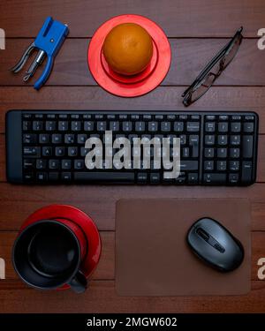 Zusammensetzung aus Tastatur, Maus auf Pad, Hefter, Brille, einer Orange und einer Tasse mit Getränk im Inneren, auf einem Holztisch Stockfoto